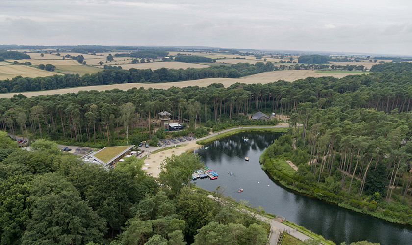 Lake hosts small paddleboats and a few moored craft, reflecting an overcast sky, while sandy beach, cabins, parking, and dense pine forest encircle it, with patchwork farmland stretching beyond.