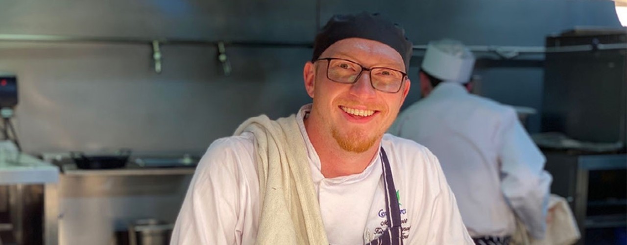 Chef smiling at Camera while standing in a kitchen looking over the hotplate pass
