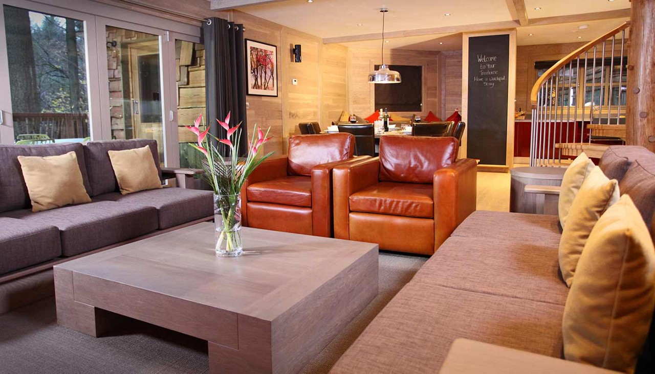 Seating group frames a low wooden coffee table holding a vase; in a warm, modern living-dining room with leather armchairs, gray sofas, large windows, spiral staircase. Text: "Welcome to the Ecurie Have a magical Stay."