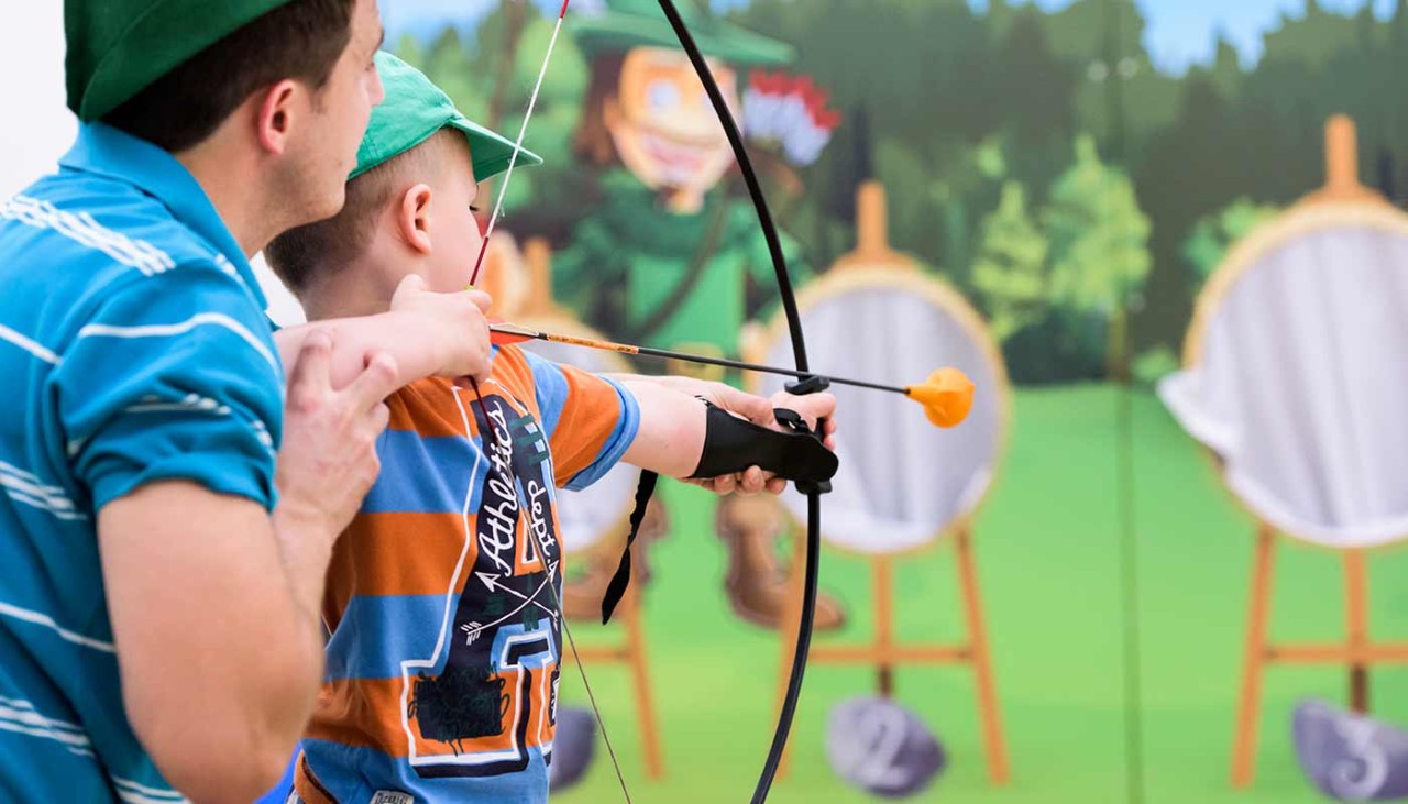 A father helps his young son aim his bow and arrow at the targets 