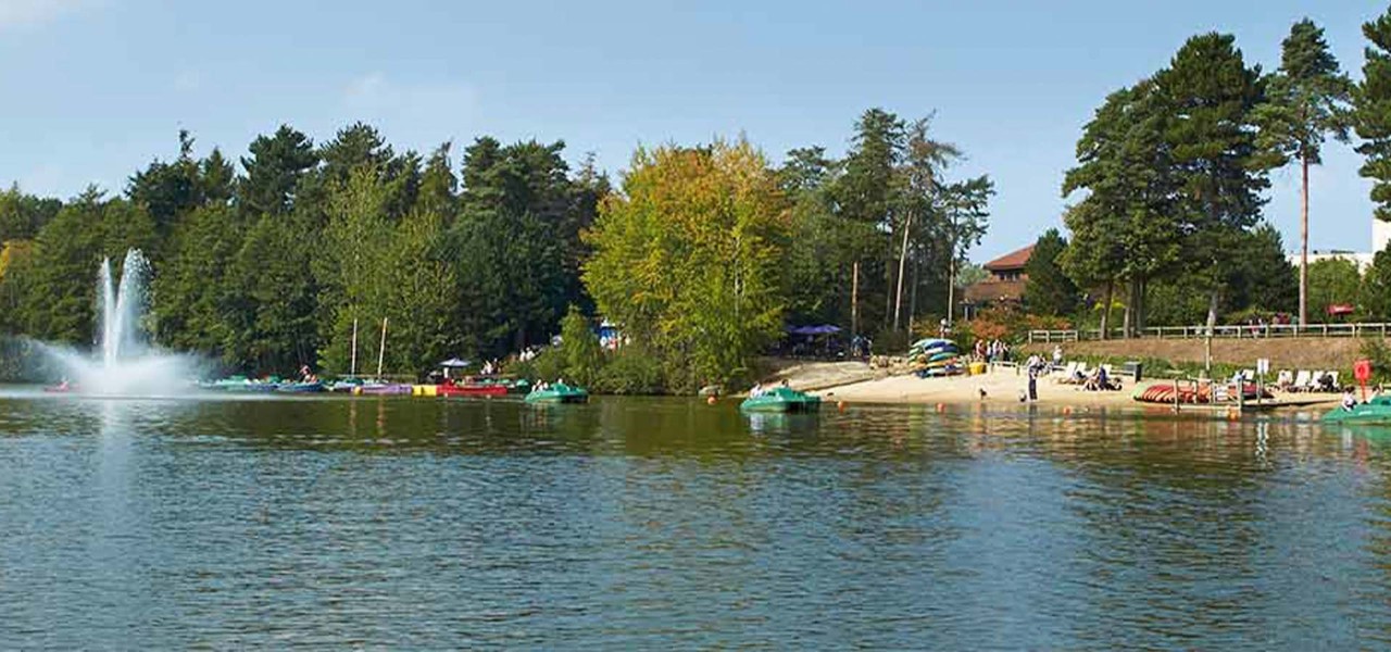 View of lake and beach with coloured boats lined up on the beach and on the lake with water fountain to the left of the lake and forest in the distance
