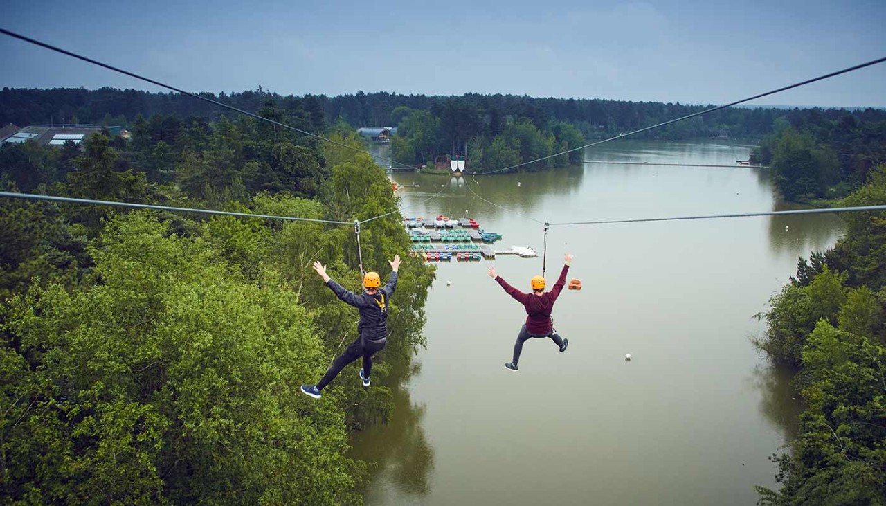 Aerial view of two men whizzing down the zip wire over the lake and forest all around them