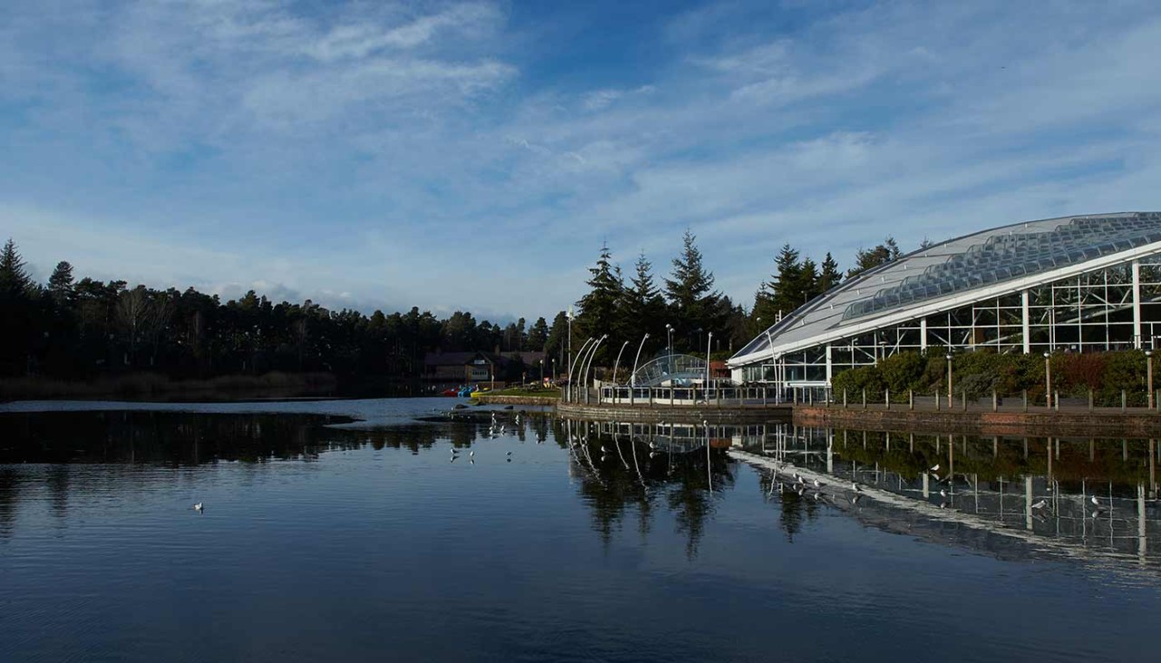 Exterior view of the side of the Subtropical Swimming Paradise with lake to the side of it and the Lakeside Inn in the distance at Whinfell Forest