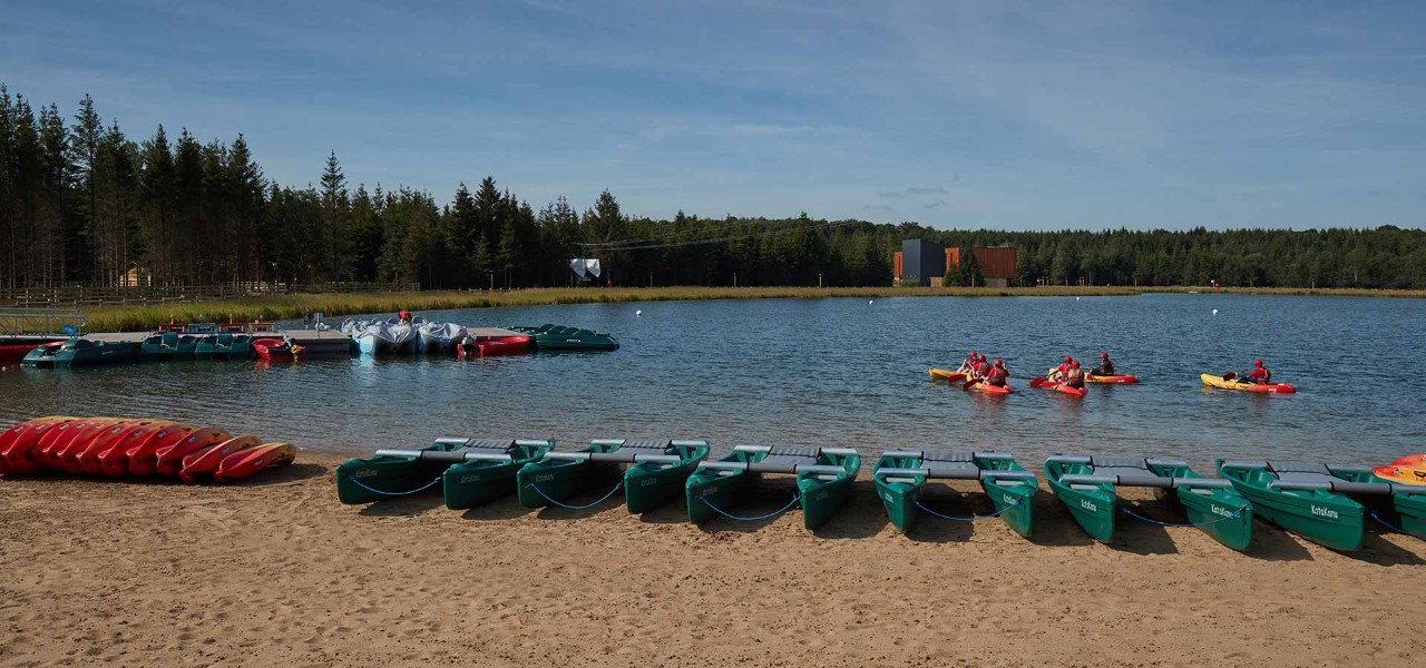 Beach at Longford Forest with boats lined up on it and some guests on boats on the lake, with forest in the distance