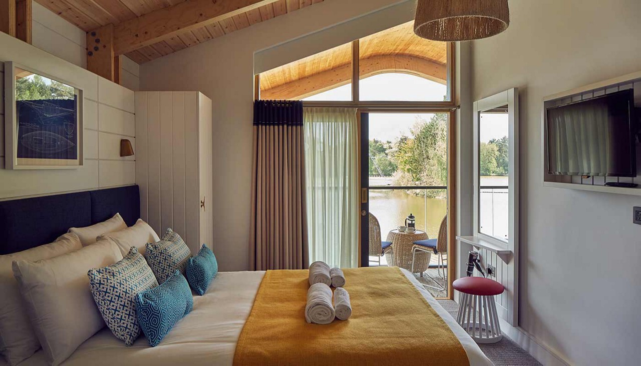Bed with yellow throw and rolled towels faces balcony doors. Outside, two chairs and a small table overlook a lake. Wood ceiling, wall TV, mirror, and curtains complete the bedroom.
