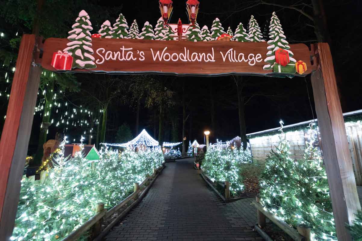 Sign with Santa's woodland village on it taken at night with trees and lights on them along a path