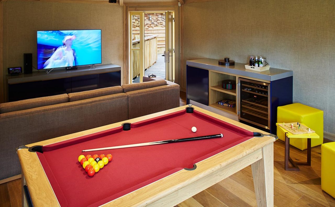 Pool table holds racked balls and cue, awaiting a break; surrounded by sofa facing TV, bar cabinet with drinks and wine fridge, yellow stools, and open doors leading to deck.