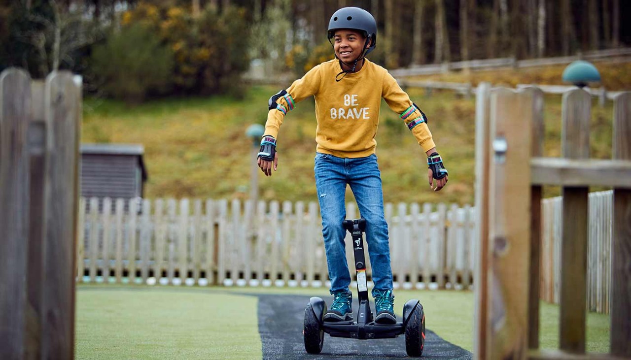 A child wearing a helmet and elbow pads rides around a track on a segway 